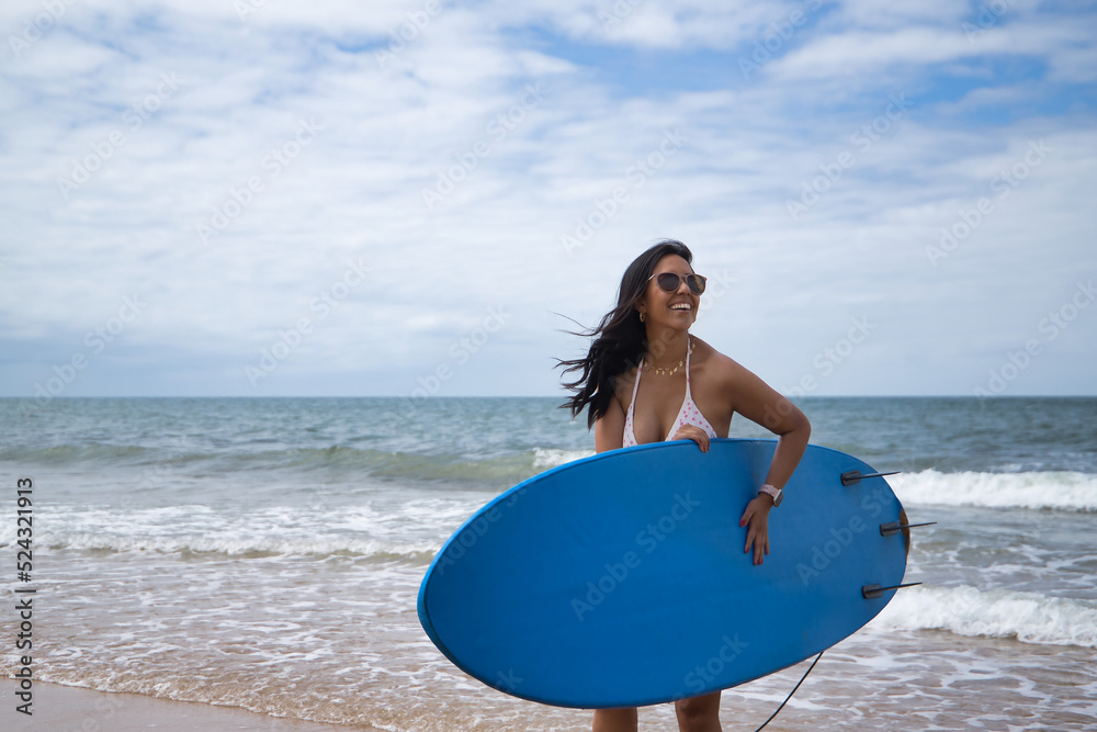 South American woman, young and beautiful, brunette with sunglasses and bikini, coming out of the water holding a blue surfboard. Concept sea, sand, sun, beach, vacation, surf, summer.