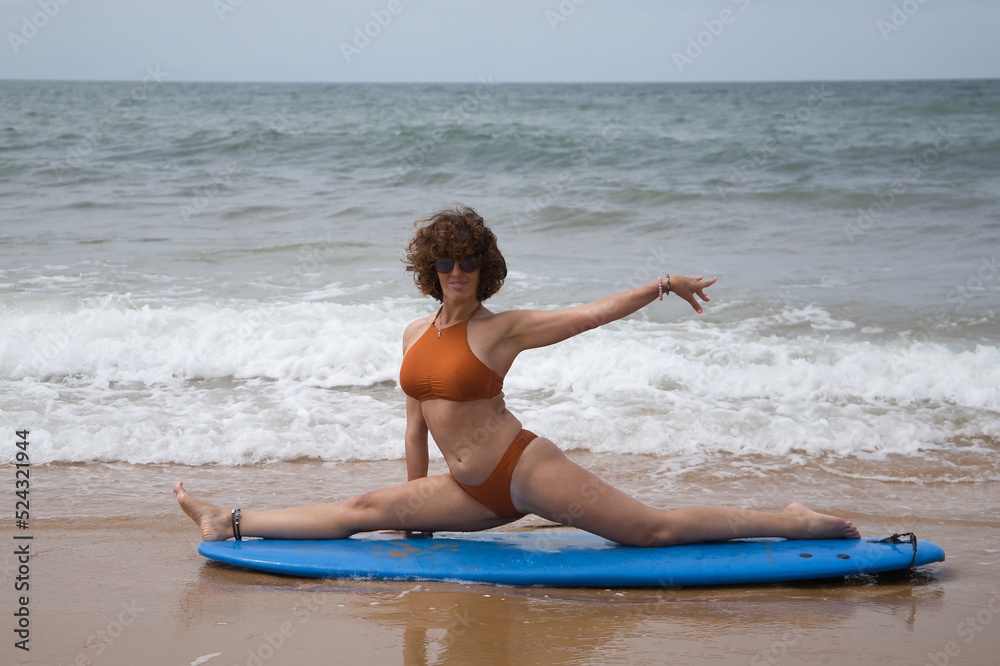 Attractive mature woman with curly hair, sunglasses and bikini, doing  ballet poses on a blue surfboard on the shore of the beach. Concept sea,  sand, sun, beach, vacation, surf, summer. Photos