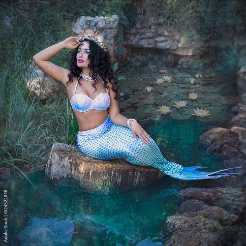 A fantastic woman, a beautiful mermaid girl is sitting on the rocks, with a beautiful hairstyle and makeup with shell ornaments. The mermaid is sitting on the shore. Fantastic concept