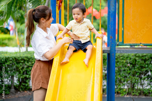 Asian mother stand and take care little boy play slide in fun park of garden and they look happy to have enjoy time together.