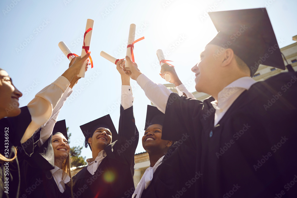 Happy diverse college or university students having fun at graduation. Joyful multiracial graduates standing together and holding up paper diploma scrolls with red ribbons. Education, success concept - obrazy, fototapety, plakaty 
