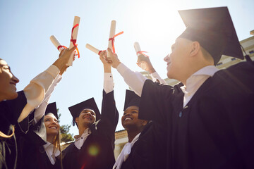 Happy diverse college or university students having fun at graduation. Joyful multiracial graduates standing together and holding up paper diploma scrolls with red ribbons. Education, success concept