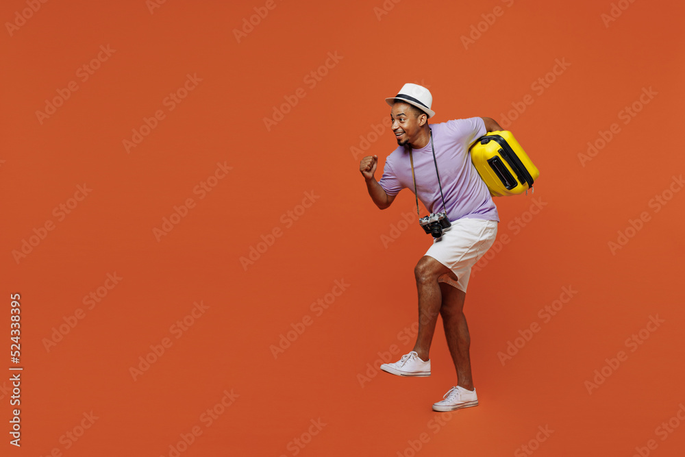 Full size traveler black man wear purple t-shirt hat go run walk with bag under armpit isolated on plain orange background. Tourist travel abroad in spare time getaway Air flight trip journey concept