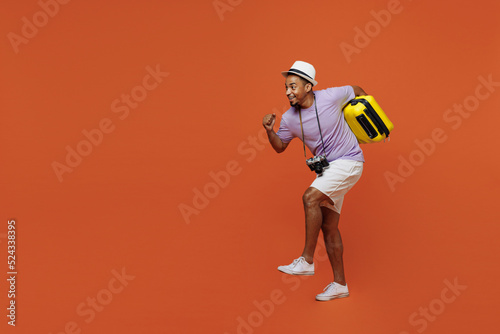 Full size traveler black man wear purple t-shirt hat go run walk with bag under armpit isolated on plain orange background. Tourist travel abroad in spare time getaway Air flight trip journey concept