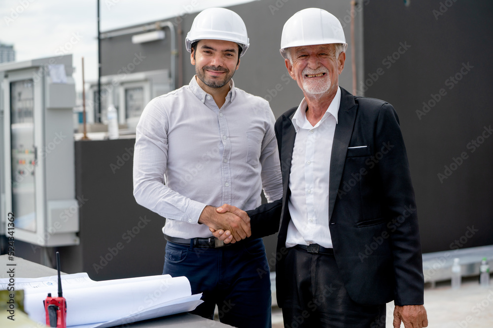 Close up young engineer shake hands with senior coworker and stay in front of line of air vent in construction site to express success of the project. Close up young engineer shake hands with senior coworker and stay in front of line of air vent in construction site to express success of the project.