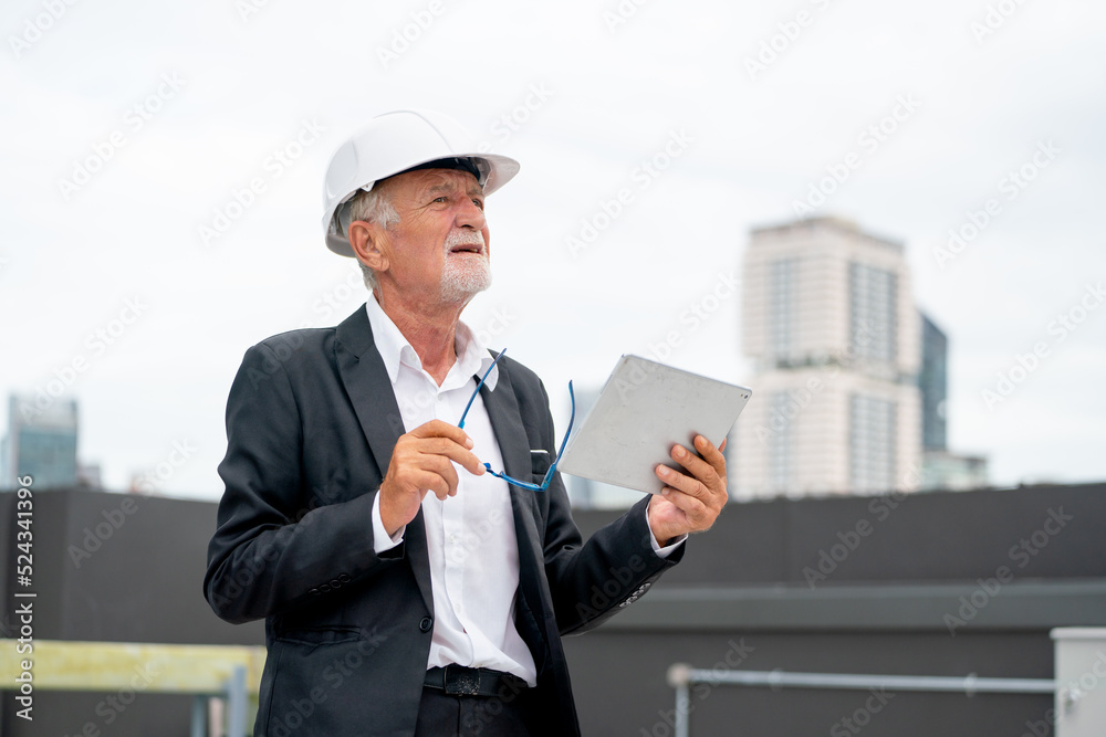 Senior engineer man using tablet to check and maintenance in area of construction site with other building as background. Senior engineer man using tablet to check and maintenance in area of construction site with other building as background.