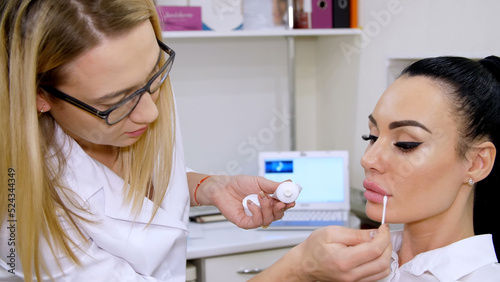 medical office  doctor puts on patient lips a cosmetic cream with a cotton swab  after injections of hyaluronic acid into lips  cleans the surface with an antiseptic. High quality photo