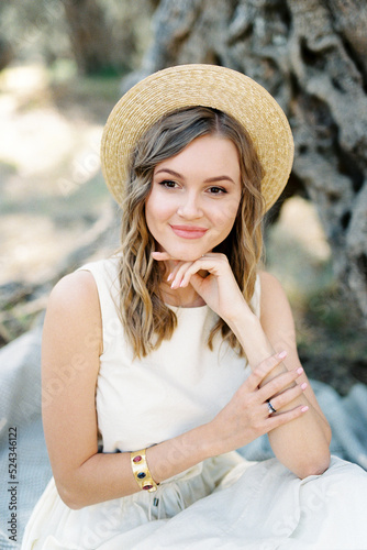 Young woman in a straw hat sits near a tree resting her chin on her hand