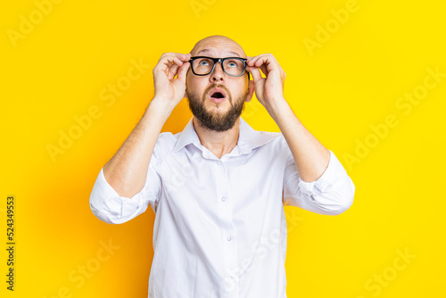 Bald young man holding glasses looks up in surprise on a yellow background