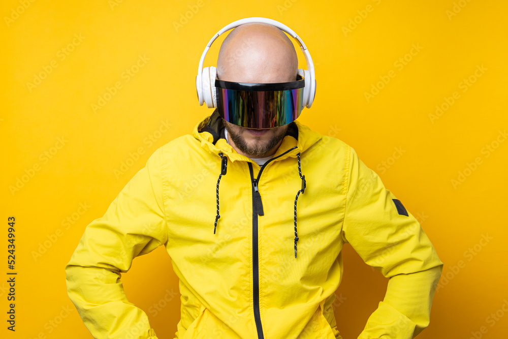 Young man in cyberpunk glasses in a yellow jacket with headphones looks down on a yellow background