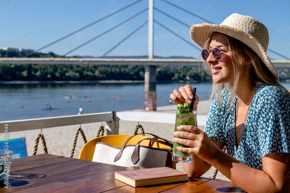 A beautiful young woman wearing a straw hat and sunglasses, having a cocktail and enjoying a lovely summer afternoon in a beach cafe by the river.