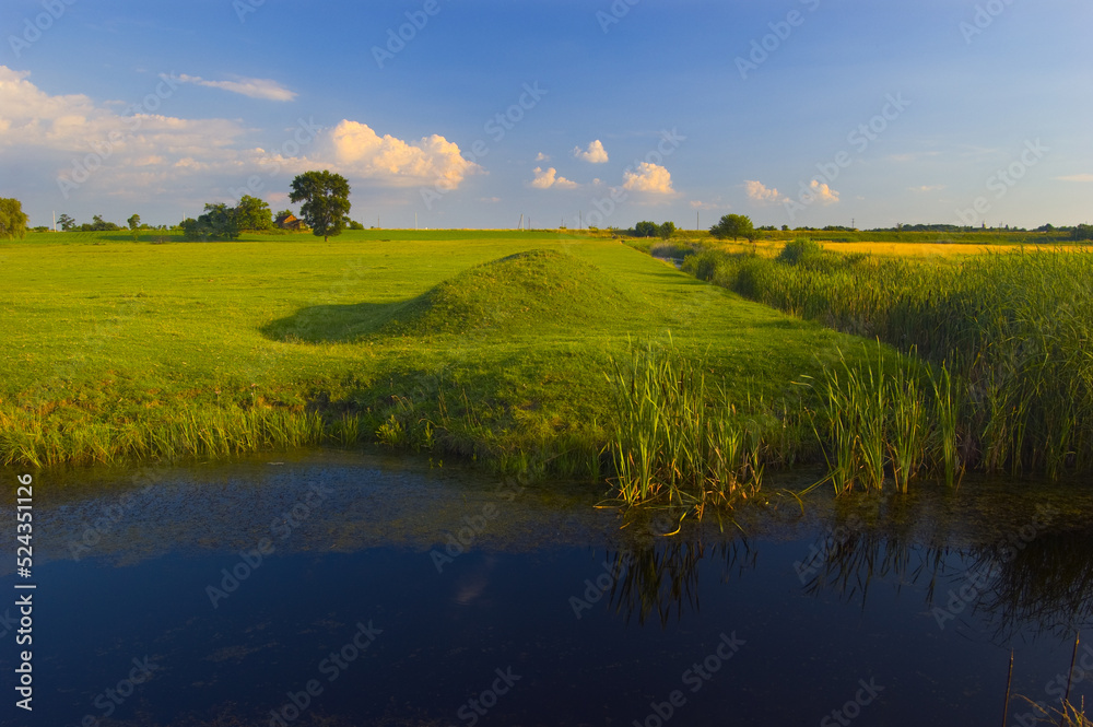 Beautiful summer landscape showing green meadow, river and blue sky with white clouds on a sunny day