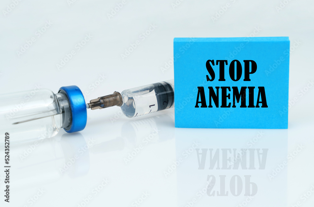 On a white reflective surface there is a syringe with an ampoule and a plate with the inscription - STOP ANEMIA On a white reflective surface there is a syringe with an ampoule and a plate with the inscription - STOP ANEMIA