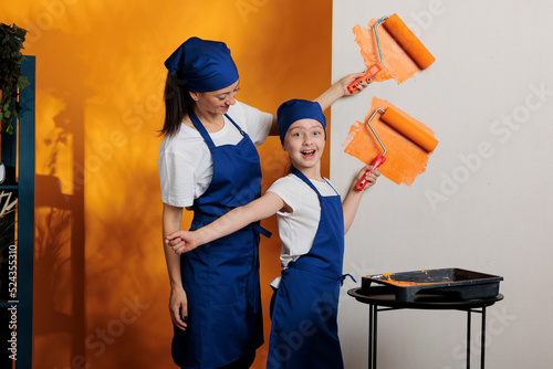 Happy people painting orange walls to redecorate house room, using renovating tools and paintbrush roller to paint color in apartment. Woman with young child dcorating home interior.