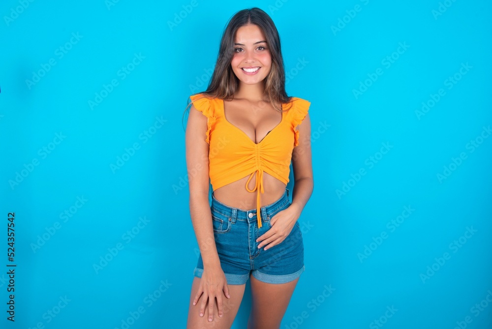 Portrait of successful beautiful brunette woman wearing orange tank top over blue background , smiling broadly with self-assured expression. Confidence and business concept.