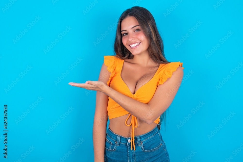 beautiful brunette woman wearing orange tank top over blue background says: wow how exciting it is, has amazed expression, shows something on blank space with palm. Advertisement concept. beautiful brunette woman wearing orange tank top over blue background says: wow how exciting it is, has amazed expression, shows something on blank space with palm. Advertisement concept.
