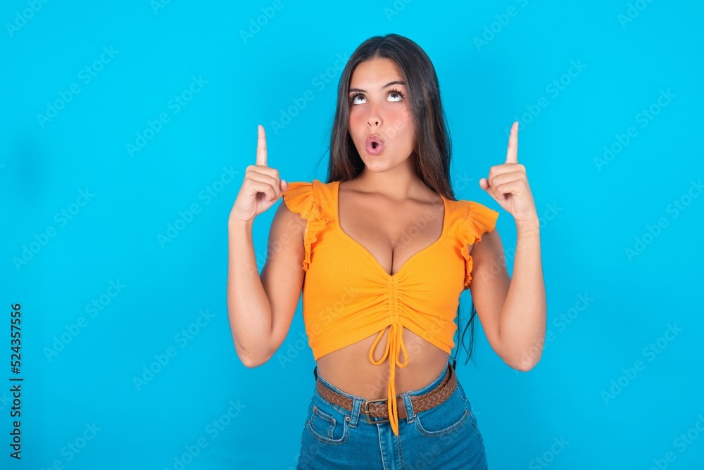 beautiful brunette woman wearing orange tank top over blue background being amazed and surprised looking and pointing up with fingers showing something strange. beautiful brunette woman wearing orange tank top over blue background being amazed and surprised looking and pointing up with fingers showing something strange.