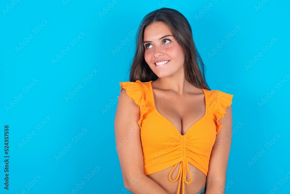 beautiful brunette woman wearing orange tank top over blue background with thoughtful expression, looks away keeps hands down bitting his lip thinks about something pleasant. beautiful brunette woman wearing orange tank top over blue background with thoughtful expression, looks away keeps hands down bitting his lip thinks about something pleasant.