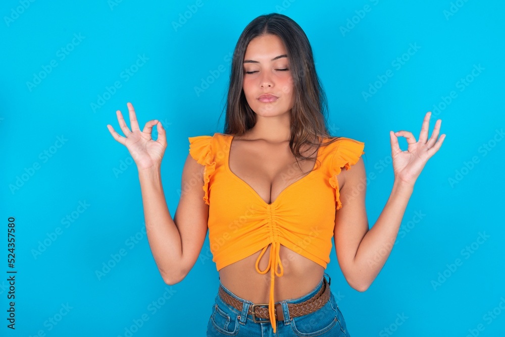 beautiful brunette woman wearing orange tank top over blue background doing yoga, keeping eyes closed, holding fingers in mudra gesture. Meditation, religion and spiritual practices. beautiful brunette woman wearing orange tank top over blue background doing yoga, keeping eyes closed, holding fingers in mudra gesture. Meditation, religion and spiritual practices.