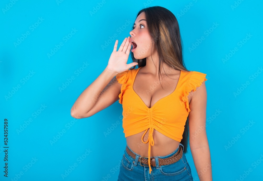beautiful brunette woman wearing orange tank top over blue background profile view, looking happy and excited, shouting and calling to copy space. beautiful brunette woman wearing orange tank top over blue background profile view, looking happy and excited, shouting and calling to copy space.