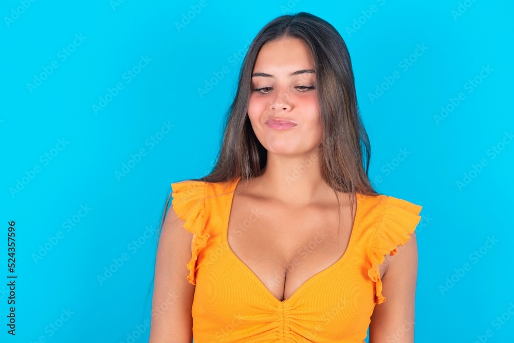 beautiful brunette woman wearing orange tank top over blue background crosses eyes, puts lips, makes grimace with awkward expression has fun alone, plays fool. beautiful brunette woman wearing orange tank top over blue background crosses eyes, puts lips, makes grimace with awkward expression has fun alone, plays fool.