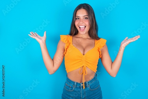 Fotografi beautiful brunette woman wearing orange tank top over blue background raising hands up, having eyes full of happiness rejoicing his great achievements