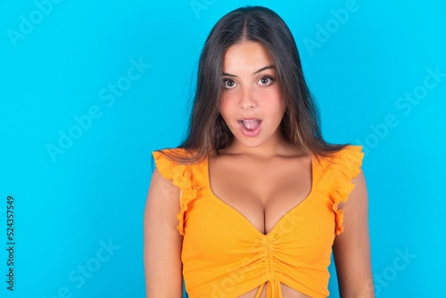 beautiful brunette woman wearing orange tank top over blue background having stunned and shocked look, with mouth open and jaw dropped exclaiming: Wow, I can't believe this Fototapeta