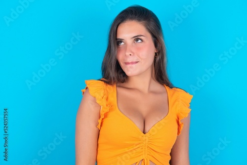 Amazed puzzled beautiful brunette woman wearing orange tank top over blue background , curves lips and has worried look, sees something awful in front.