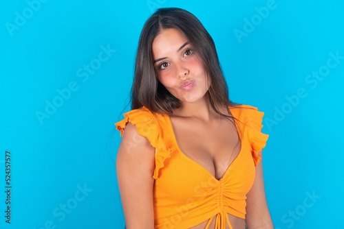 Canvas Print beautiful brunette woman wearing orange tank top over blue background , keeps lips as going to kiss someone, has glad expression, grimace face