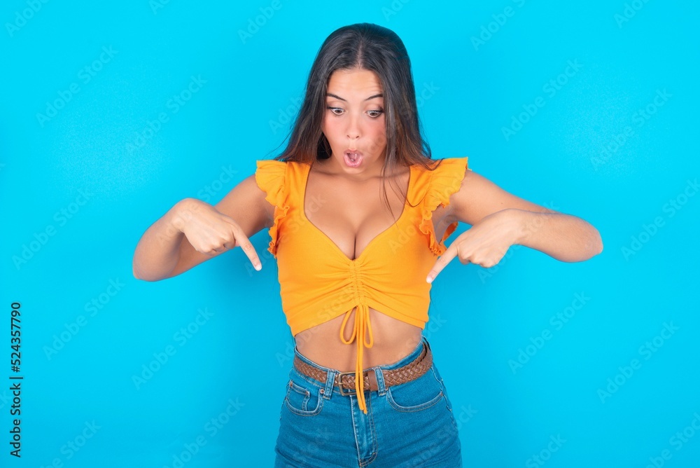 Amazed brunette woman wearing orange tank top over blue background points down with fore fingers, opens mouth being shocked. Advertisement concept. Amazed brunette woman wearing orange tank top over blue background points down with fore fingers, opens mouth being shocked. Advertisement concept.