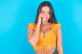 brunette woman wearing orange tank top over blue background  excited looking to the side hand on face. Advertisement and amazement concept.