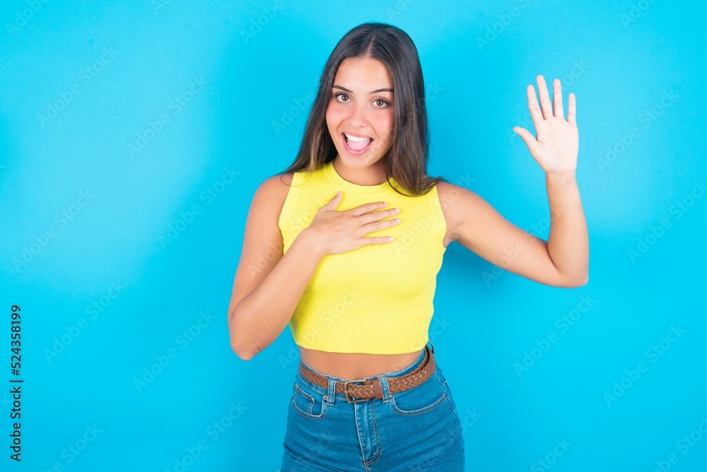 I swear, promise you not regret. Portrait of sincere brunette woman wearing yellow tank top over blue wall raising one arm and hold hand on heart as give oath, telling truth, want you to believe. I swear, promise you not regret. Portrait of sincere brunette woman wearing yellow tank top over blue wall raising one arm and hold hand on heart as give oath, telling truth, want you to believe.