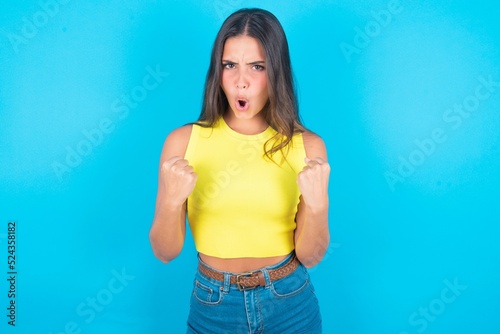 Joyful excited lucky brunette woman wearing yellow tank top over blue background cheering, celebrating success, screaming yes with clenched fists