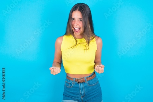 brunette woman wearing yellow tank top over blue background raising fists up screaming with joy being happy to achieve goals.