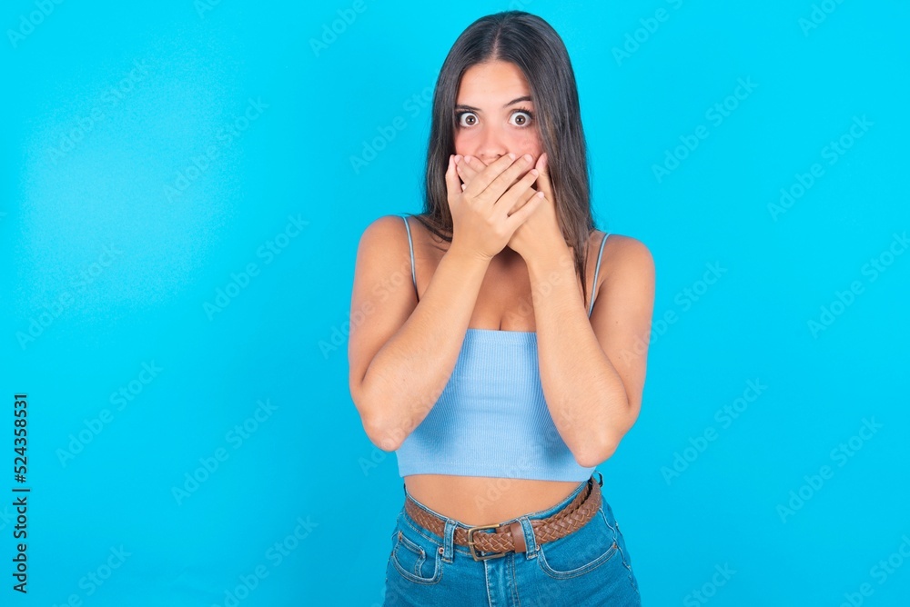 Stunned beautiful brunette woman wearing blue tank top over blue background covers mouth with both hands being afraid from something or after hearing stunning gossips. Stunned beautiful brunette woman wearing blue tank top over blue background covers mouth with both hands being afraid from something or after hearing stunning gossips.