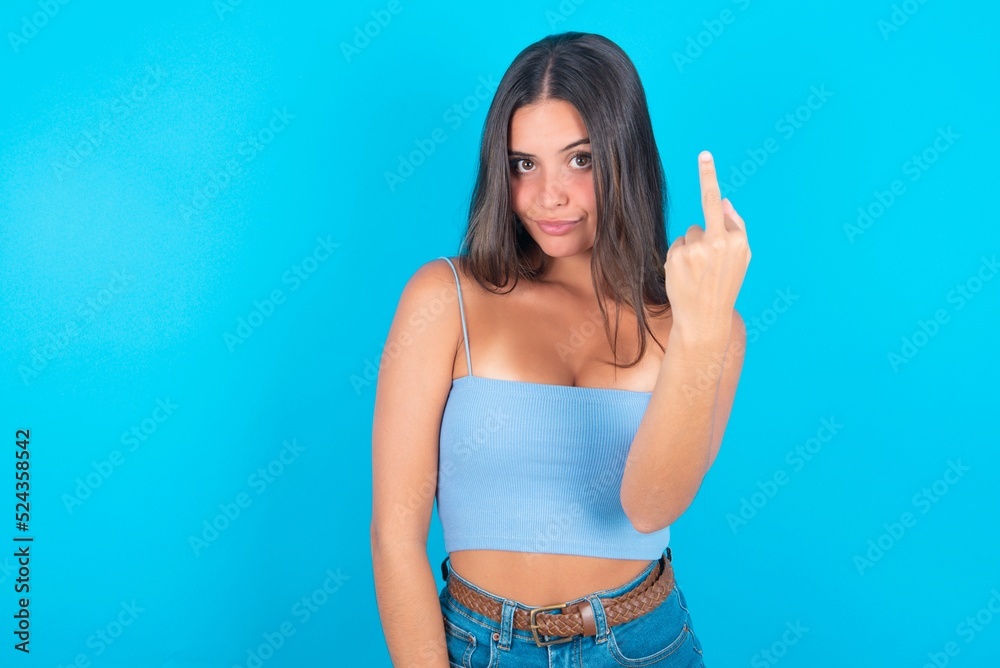 beautiful brunette woman wearing blue tank top over blue background shows middle finger bad sign asks not to bother. Provocation and rude attitude. beautiful brunette woman wearing blue tank top over blue background shows middle finger bad sign asks not to bother. Provocation and rude attitude.