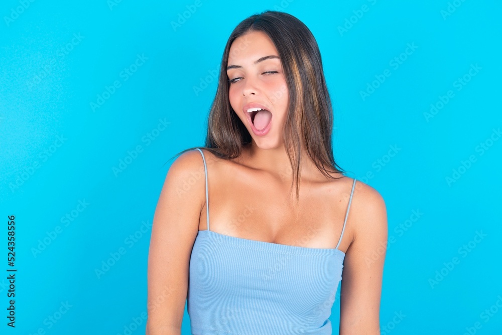beautiful brunette woman wearing blue tank top over blue background yawns with opened mouth stands. Daily morning routine beautiful brunette woman wearing blue tank top over blue background yawns with opened mouth stands. Daily morning routine