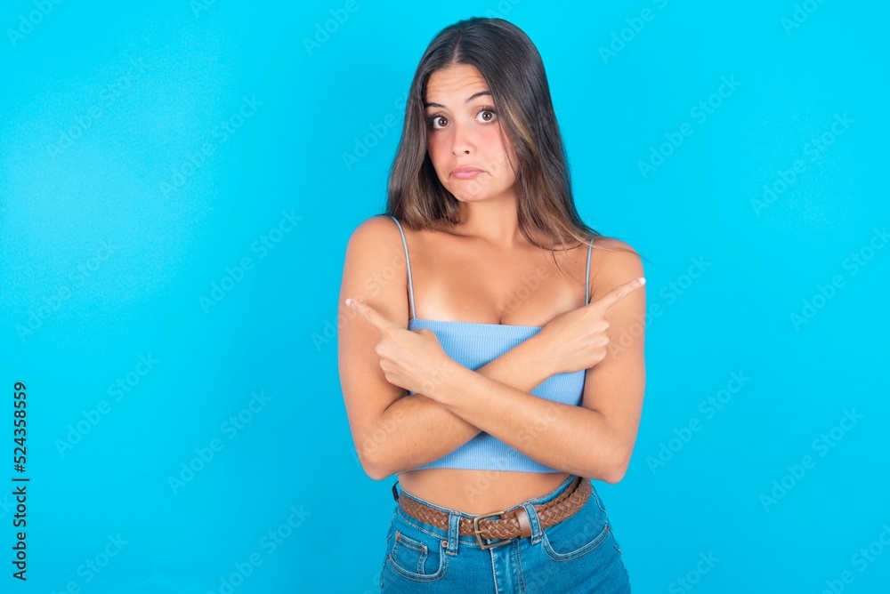 Fototapeta premium beautiful brunette woman wearing blue tank top over blue background crosses arms and points at different sides hesitates between two items or variants. Needs help with decision
