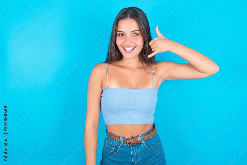 beautiful brunette woman wearing blue tank top over blue background makes phone gesture, says call me back again, has glad expression. beautiful brunette woman wearing blue tank top over blue background makes phone gesture, says call me back again, has glad expression.