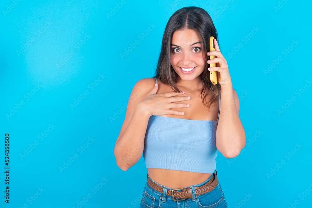 Smiling beautiful brunette woman wearing blue tank top over blue background talks via cellphone, enjoys pleasant great conversation. People, technology, communication concept Smiling beautiful brunette woman wearing blue tank top over blue background talks via cellphone, enjoys pleasant great conversation. People, technology, communication concept