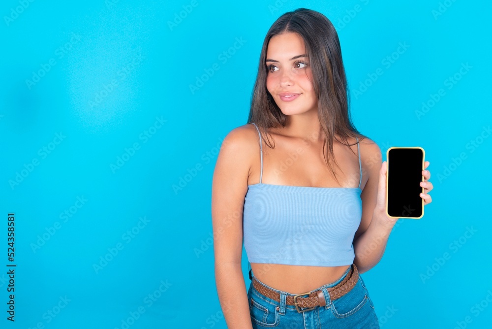 beautiful brunette woman wearing blue tank top over blue background holds new mobile phone and looks mysterious aside shows blank display of modern cellular beautiful brunette woman wearing blue tank top over blue background holds new mobile phone and looks mysterious aside shows blank display of modern cellular