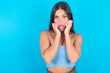 Speechless beautiful brunette woman wearing blue tank top over blue background  keeps hands near opened mouth reacts to shocking news stares wondered at camera