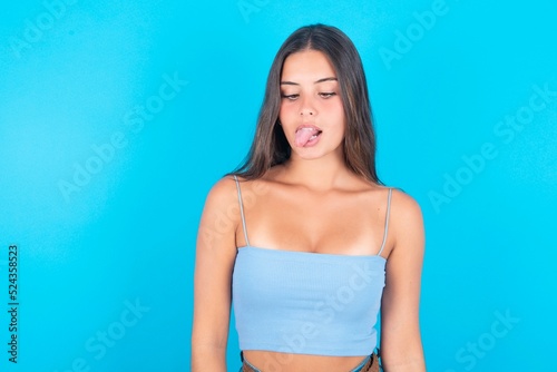 Funny beautiful brunette woman wearing blue tank top over blue background makes grimace and crosses eyes plays fool has fun alone sticks out tongue Fototapet
