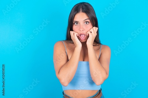 Speechless beautiful brunette woman wearing blue tank top over blue background keeps hands near opened mouth reacts to shocking news stares wondered at camera