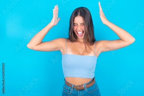 beautiful brunette woman wearing blue tank top over blue background goes crazy as head goes around feels stressed because of horrible situation