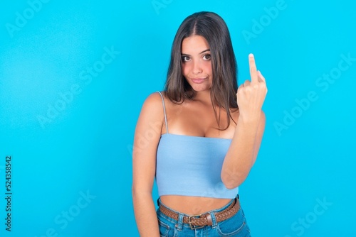 beautiful brunette woman wearing blue tank top over blue background shows middle finger bad sign asks not to bother. Provocation and rude attitude. photo