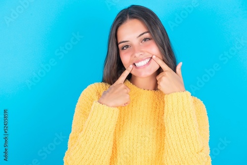 Happy beautiful brunette woman wearing yellow sweater over blue background with toothy smile, keeps index fingers near mouth, fingers pointing and forcing cheerful smile photo