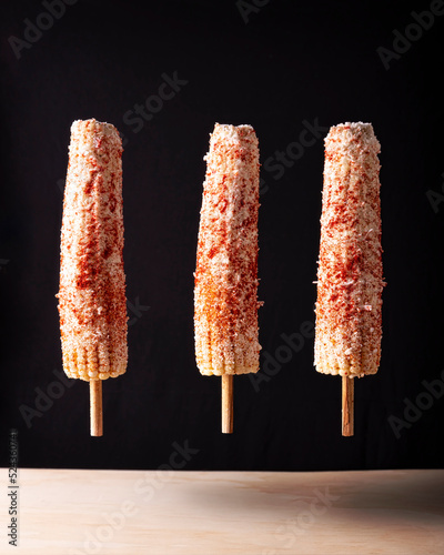 Elotes Callejeros are a popular street snack in Mexico, it is tender corn cooked with spices, skewered on a wooden stick covered with mayonnaise or sour cream, fresh cheese and piquin chili powder. photo