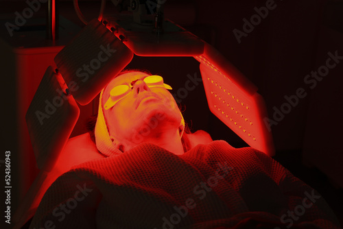 Express facial treatment with led therapy. Beautiful girl on a light therapy procedure. LED lamp with red and yellow light. Safe skin care. Woman in protective glasses. Beauty and wellness concept.  photo