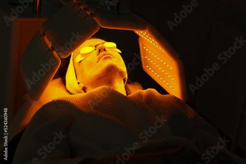 Express facial treatment with led therapy. Beautiful girl on a light therapy procedure. LED lamp with red and yellow light. Safe skin care. Woman in protective glasses. Beauty and wellness concept. 